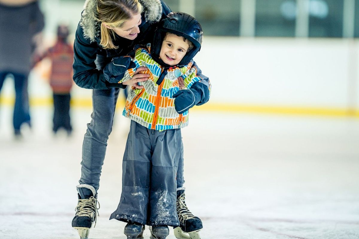 A woman helps a young child ice skate, both smiling, with the child wearing a helmet and colorful jacket on an indoor ice rink.
