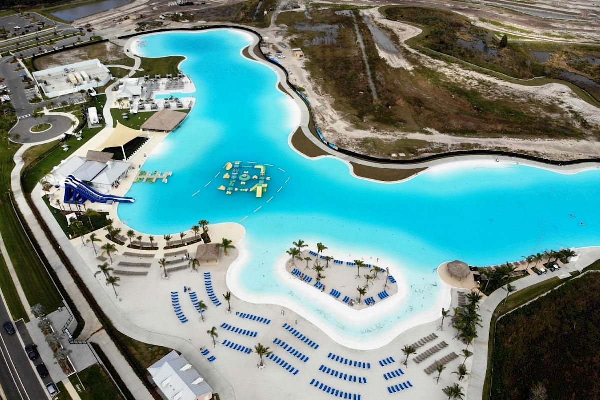 The image shows a large, lagoon-style swimming pool with water slides, floating inflatables, and numerous beach chairs. The area is surrounded by sandy beach.