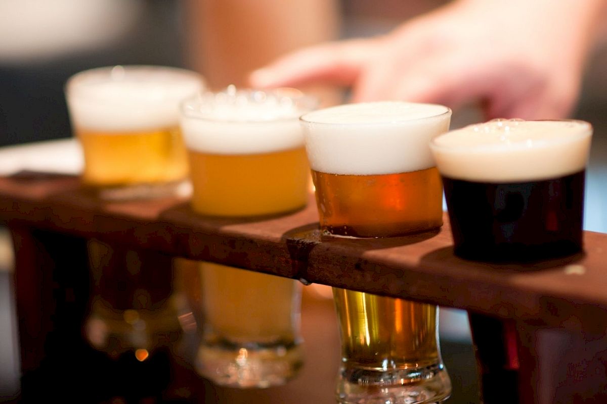 A wooden tray holds four different types of beer in small glasses, with varying colors and foam levels, while a hand reaches towards them.