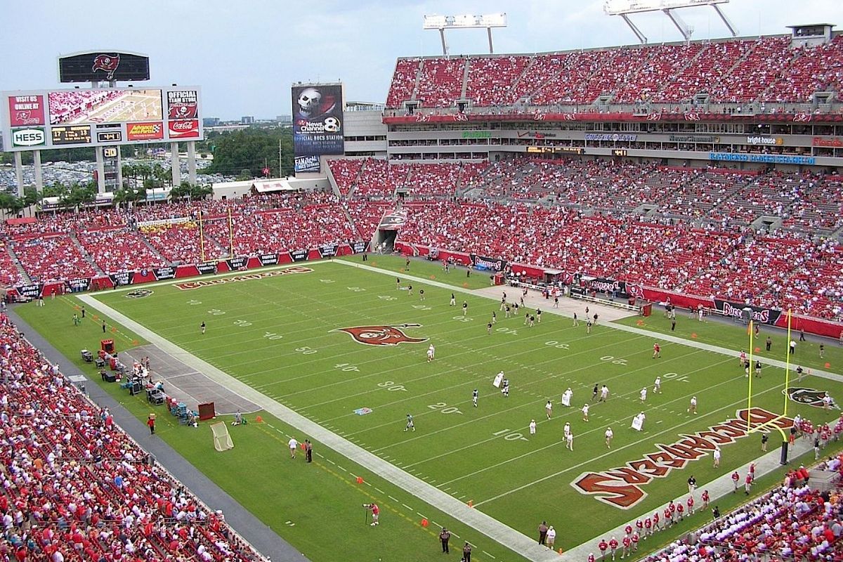 A packed stadium with a football game in progress, featuring red and white-clad players. There is a large scoreboard and many spectators.
