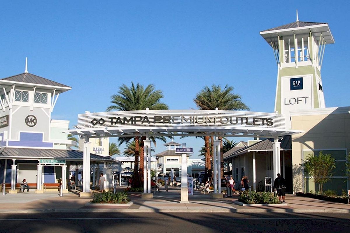 The image shows the entrance to Tampa Premium Outlets, featuring a sign, palm trees, and stores with the 'LOFT' building visible in the background.