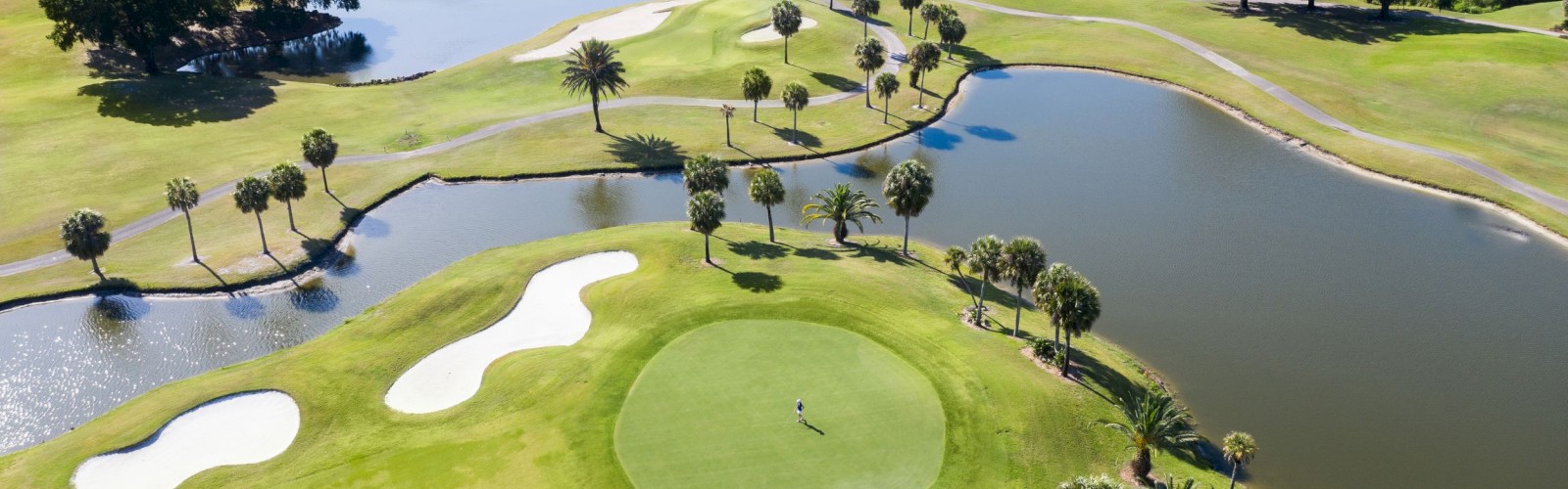 An aerial view of a golf course featuring a putting green, sand traps, ponds, and palm trees, with a lone golfer on the green.