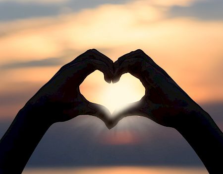 Silhouetted hands forming a heart shape against a sunset or sunrise background, with light shining through the heart opening.
