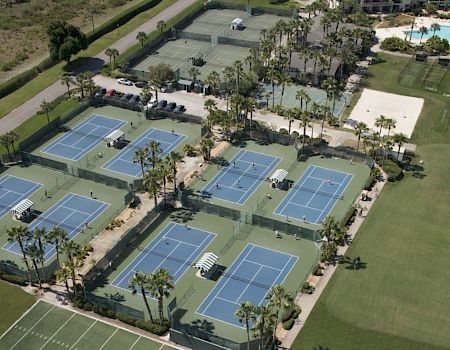 The image shows an aerial view of a sports complex with multiple tennis courts, a pool area, and adjacent green spaces near a water body, ending the sentence.