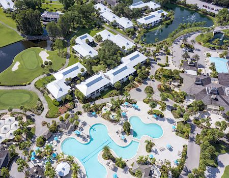 An aerial view of a resort with large pools, white-roofed buildings, greenery, and water bodies. The scene includes a golf course.