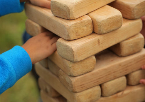 Several hands are playing with a large Jenga tower made of wooden blocks outdoors, focused on balancing the structure.