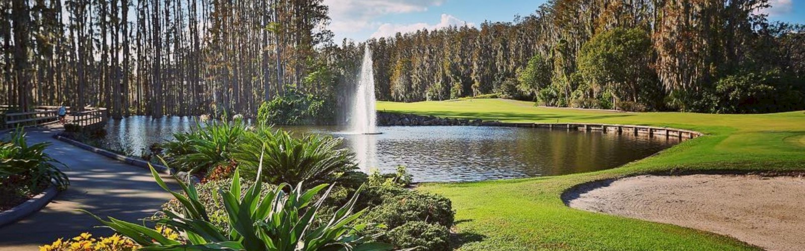 A serene golf course features a lake with a central fountain, surrounded by lush greenery, colorful plants, a bridge, and a sand trap.