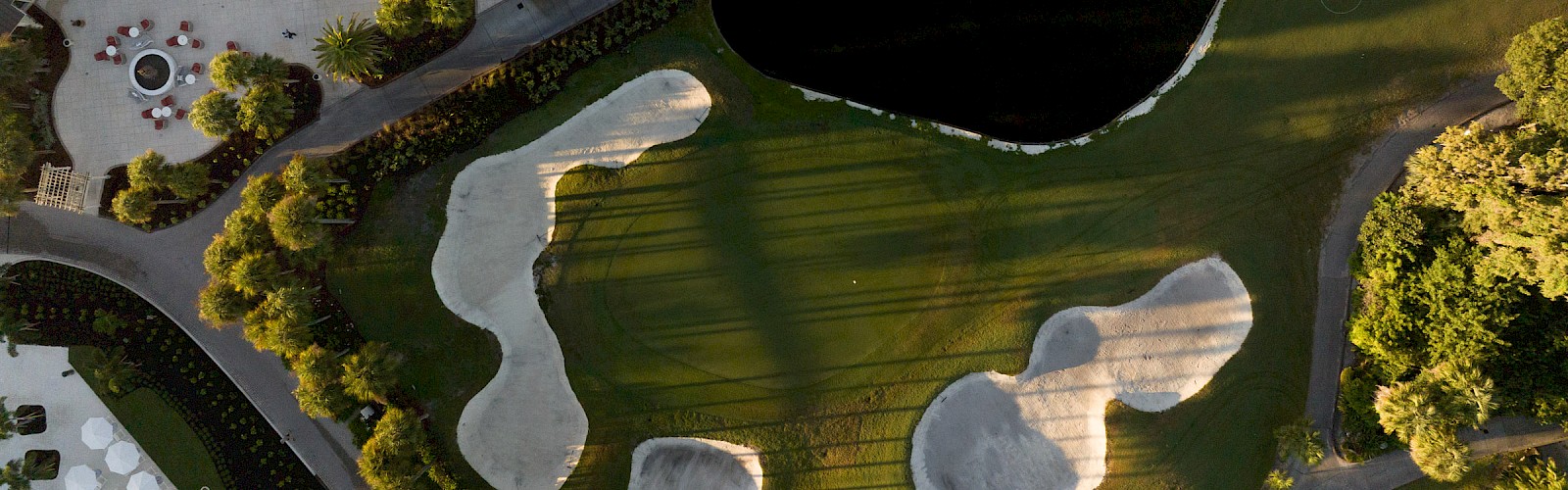 Aerial view of a golf course with greens, sand bunkers, water hazard, and surrounding trees; a curved cart path winds beside the fairways.