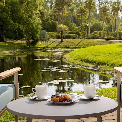 Two chairs overlook a pond, with food and coffee on a table, surrounded by lush greenery and trees.