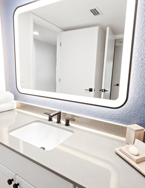 A modern bathroom with a lit mirror, single sink, neatly folded towels, and toiletries on the counter, featuring a sleek design.
