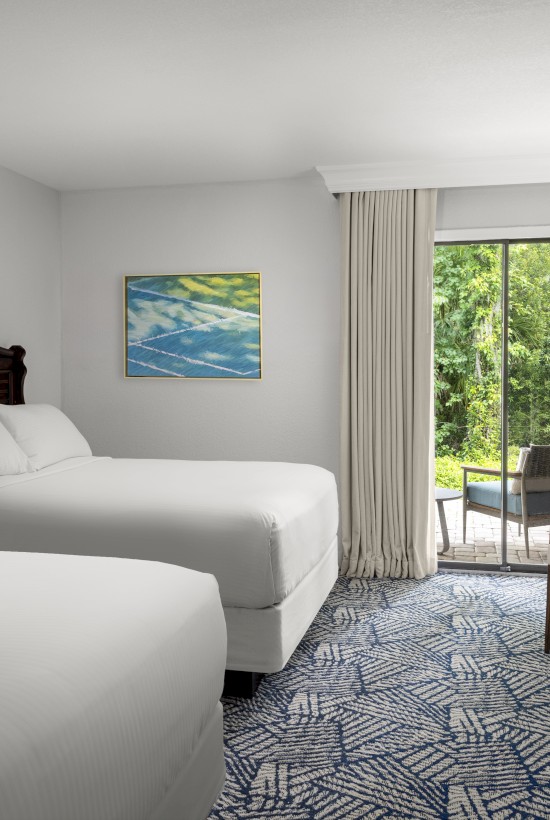 A hotel room with two beds, a large window, TV, lamp, artwork, chair, and outdoor view of greenery through glass doors.