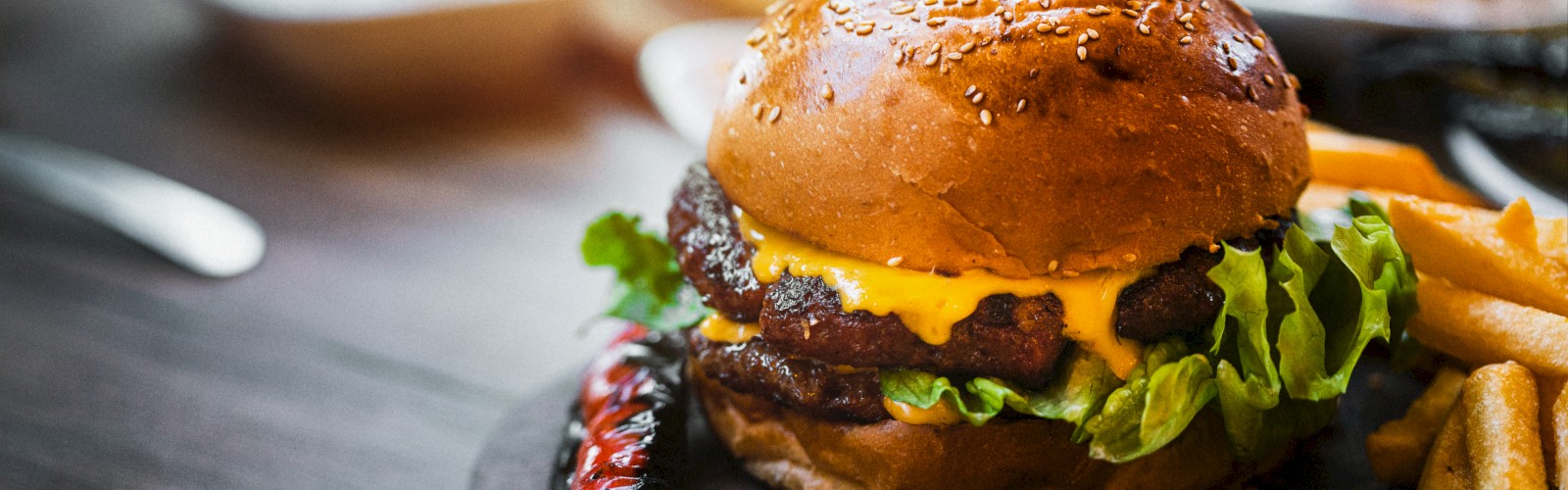 A cheeseburger with lettuce and sauce sits on a wooden board, accompanied by fries and a pepper, with sauces in the background.