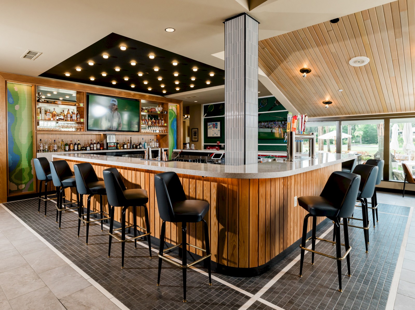 A modern bar with black chairs, wooden accents, a TV, and outdoor seating visible through windows in a well-lit space.