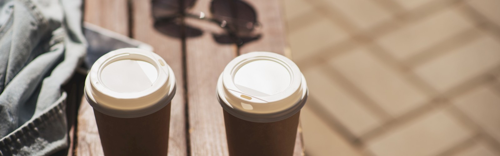 Two coffee cups on a bench with sunglasses and a piece of clothing nearby, on a sunny day.