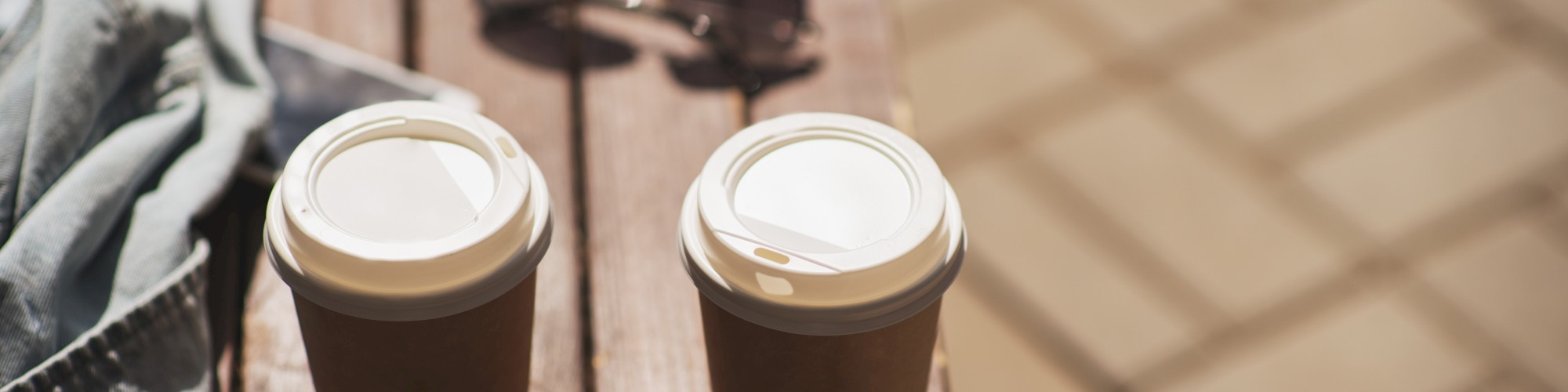 Two coffee cups on a bench with sunglasses and a piece of clothing nearby, on a sunny day.