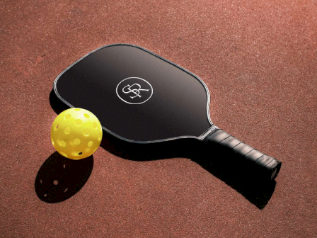 A black pickleball paddle with a logo is next to a yellow perforated ball on a textured surface.