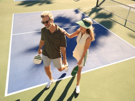 Two people on a pickleball court, holding paddles. One person is stretching, while the other places a hand on their shoulder.