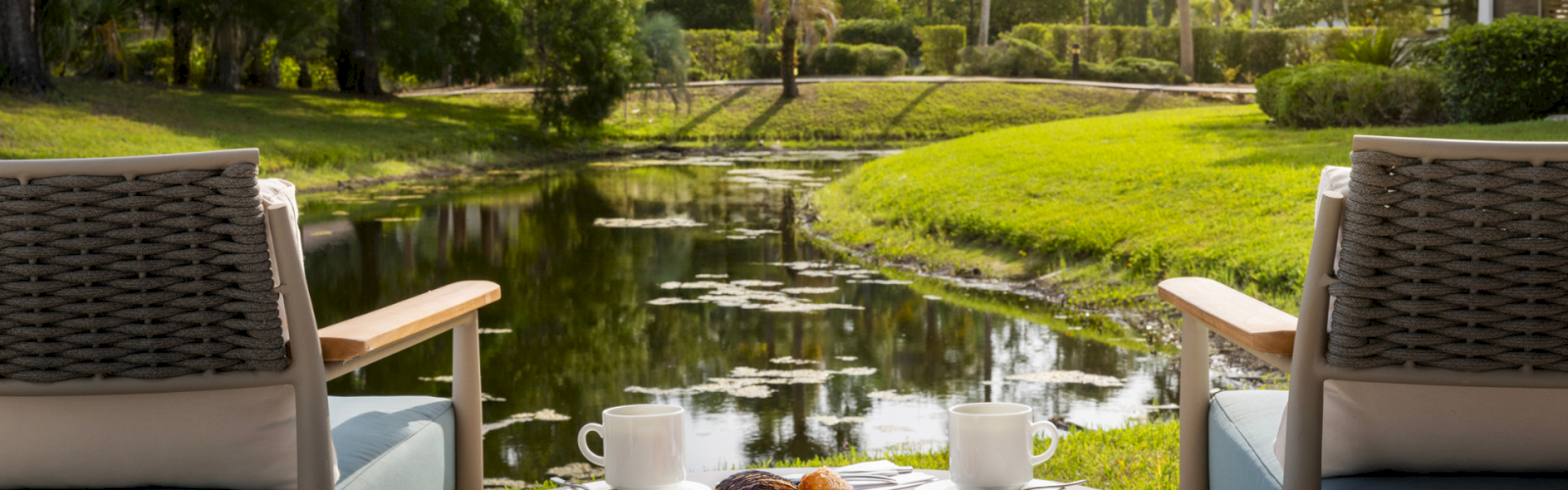 A peaceful outdoor breakfast scene with two chairs, a small table, coffee, pastries, and a scenic view of a pond surrounded by trees.