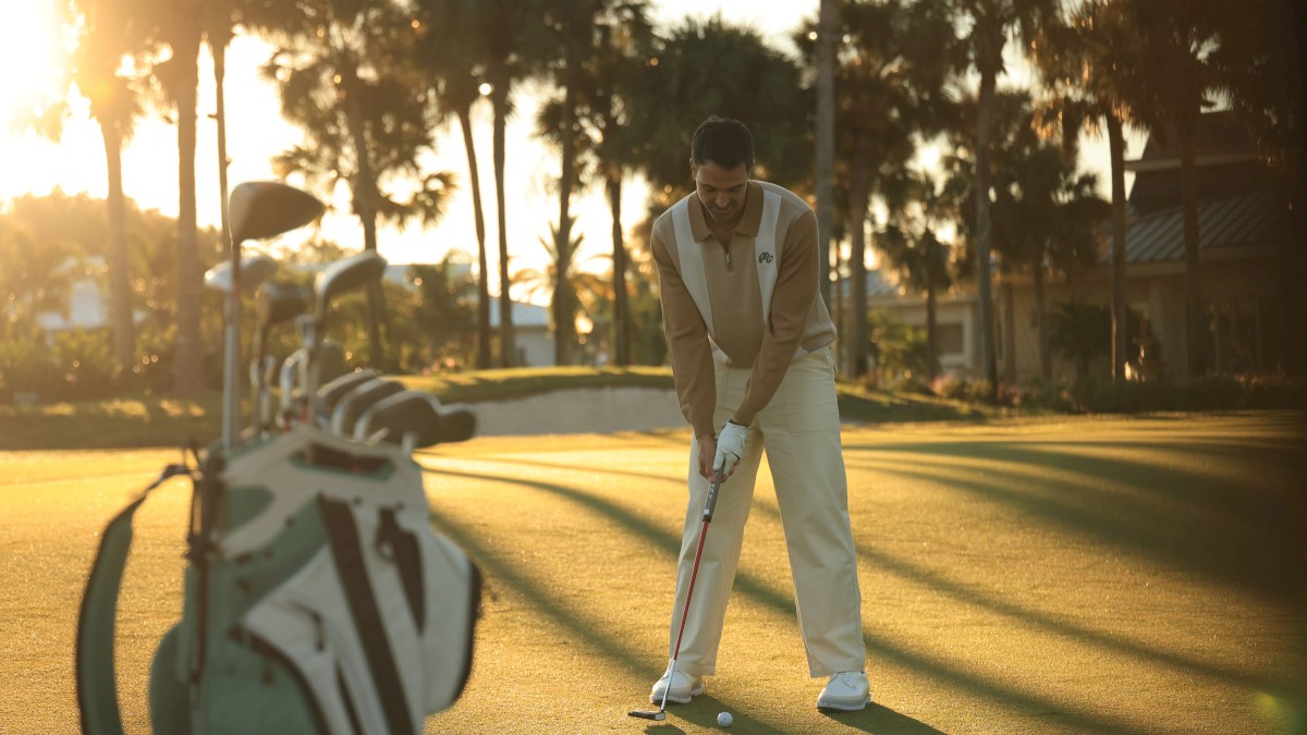 A man practicing golf on a sunny course during sunset with a golf bag nearby.