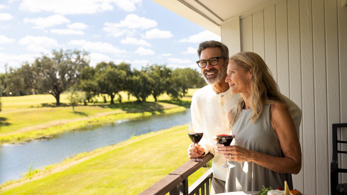 An older man and woman enjoy wine on a balcony overlooking a lake on a sunny day.