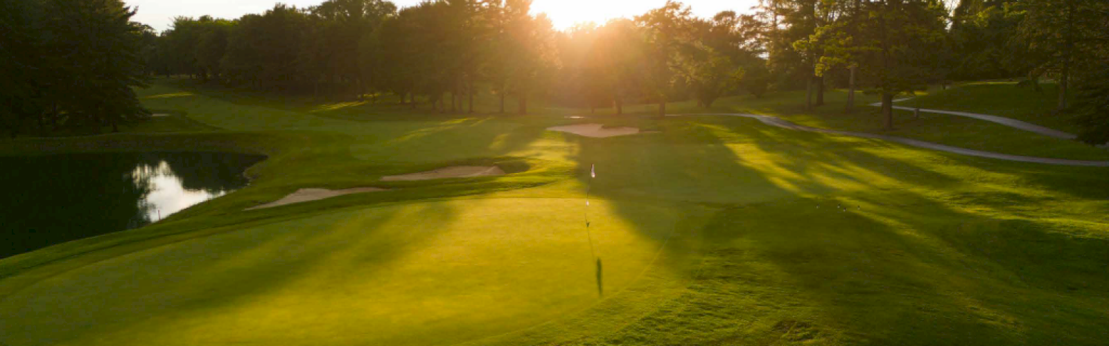 A golf course during sunset, with lush green fairways, a flagstick, sand traps, trees, and sunlight filtering through.