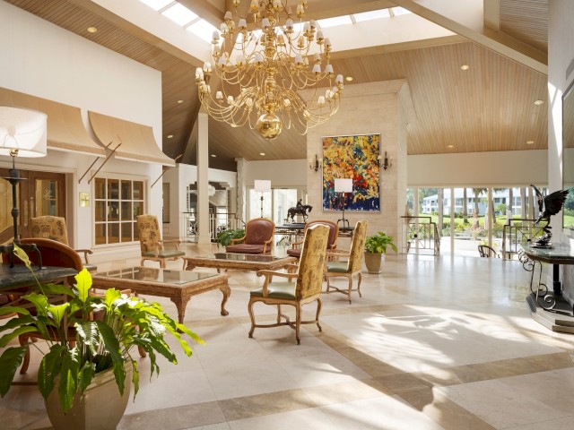 An elegant, spacious lobby area with a high ceiling, chandelier, decorative furniture, plants, large windows, and a colorful abstract painting.