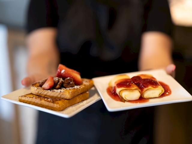The image shows a person holding two plates of food: one with French toast topped with strawberries and the other with crepes drizzled with strawberry sauce.