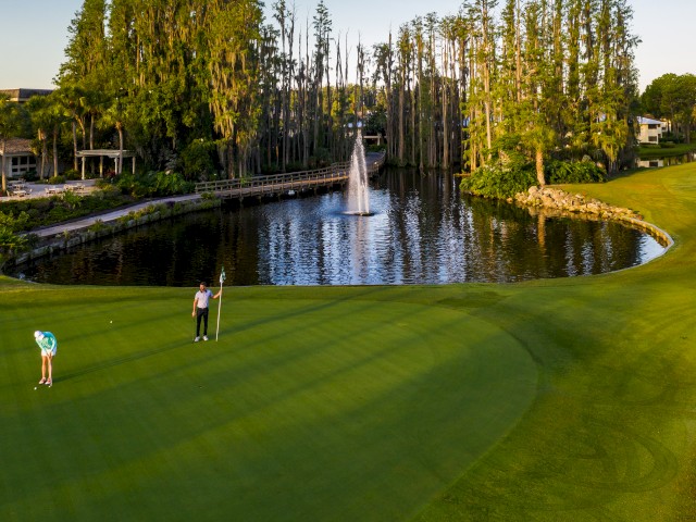 Two people on a golf course with a pond and fountain, surrounded by trees and greenery, with one lining up a putt.