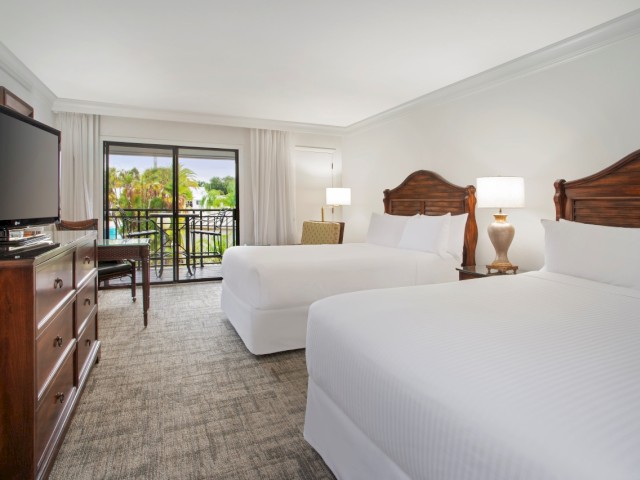 A hotel room with two white beds, a TV on a dresser, a desk, two lamps, and a balcony with a view of greenery outside.