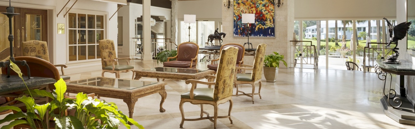 A spacious, elegant lobby with luxurious chandeliers, plush seating, artwork, and large windows letting in natural sunlight, featuring potted plants.