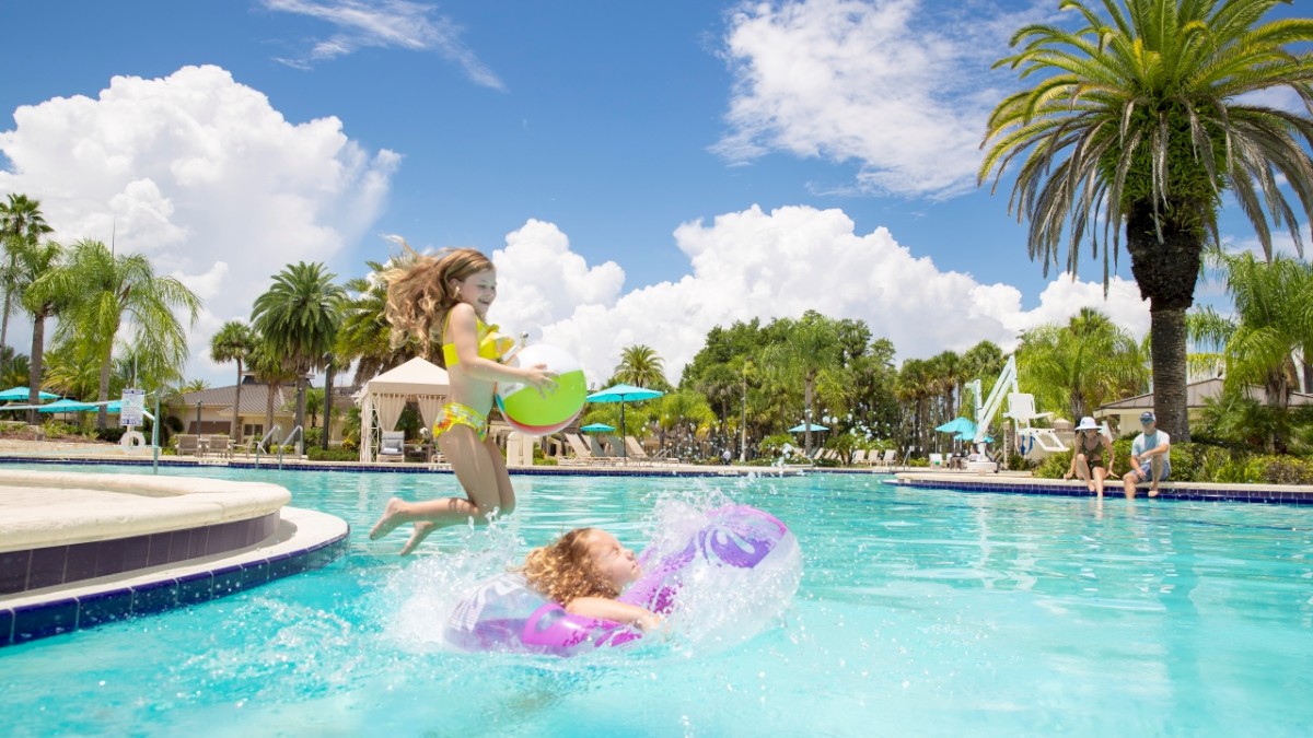 Two children are playing in a sunny swimming pool surrounded by palm trees; one child is jumping in while holding a green ring float.
