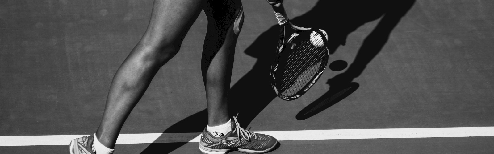 A person playing tennis, captured in black and white, mid-action with the shadow prominently visible on the court surface.