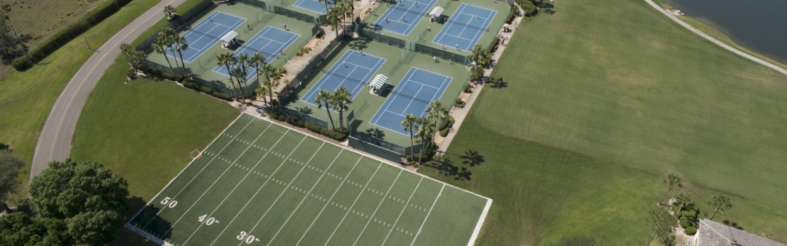 An aerial view of a recreational facility featuring tennis courts, a football field, a swimming pool, landscaped areas, and nearby water bodies.