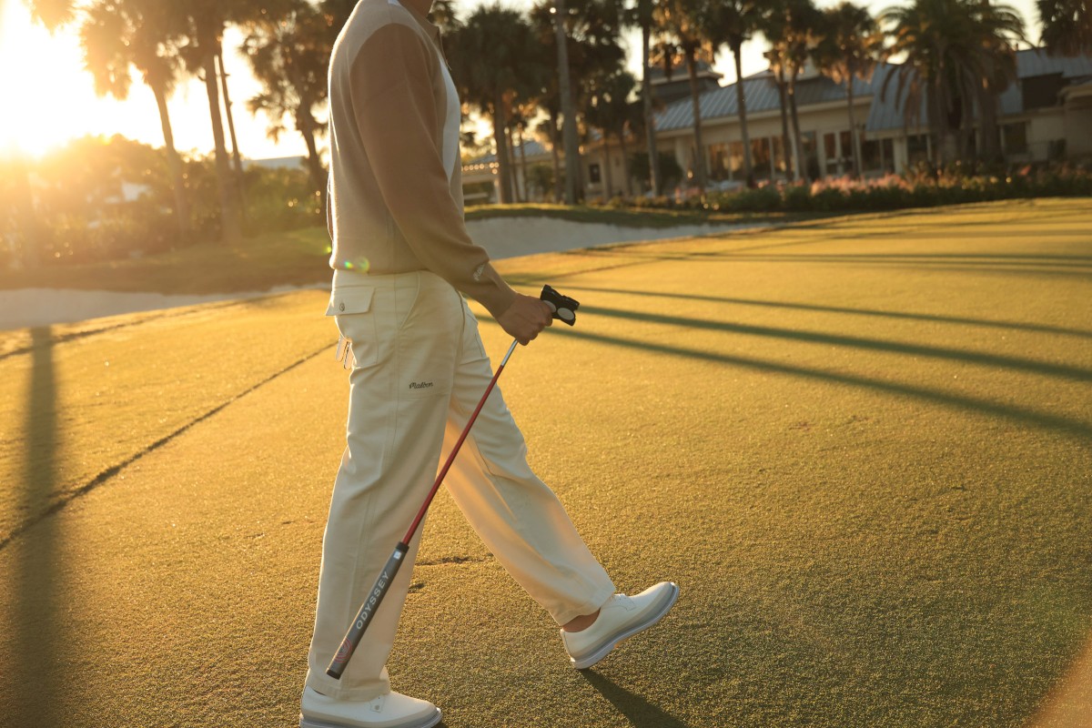 A person in light clothing walks on a golf course at sunset, holding two golf clubs, with shadows extending across the grass.