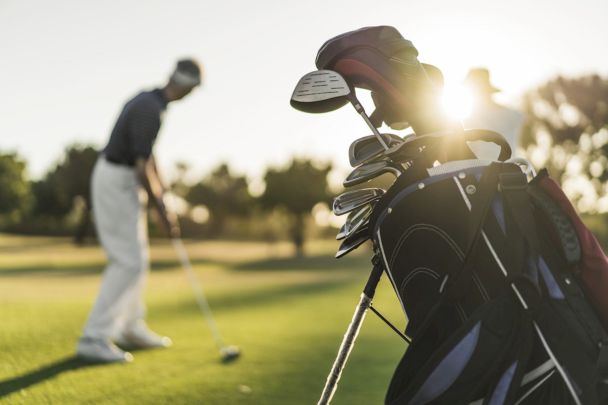 A golfer on a course is preparing to putt, while a bag of golf clubs is visible in the foreground with sunlight streaming through.