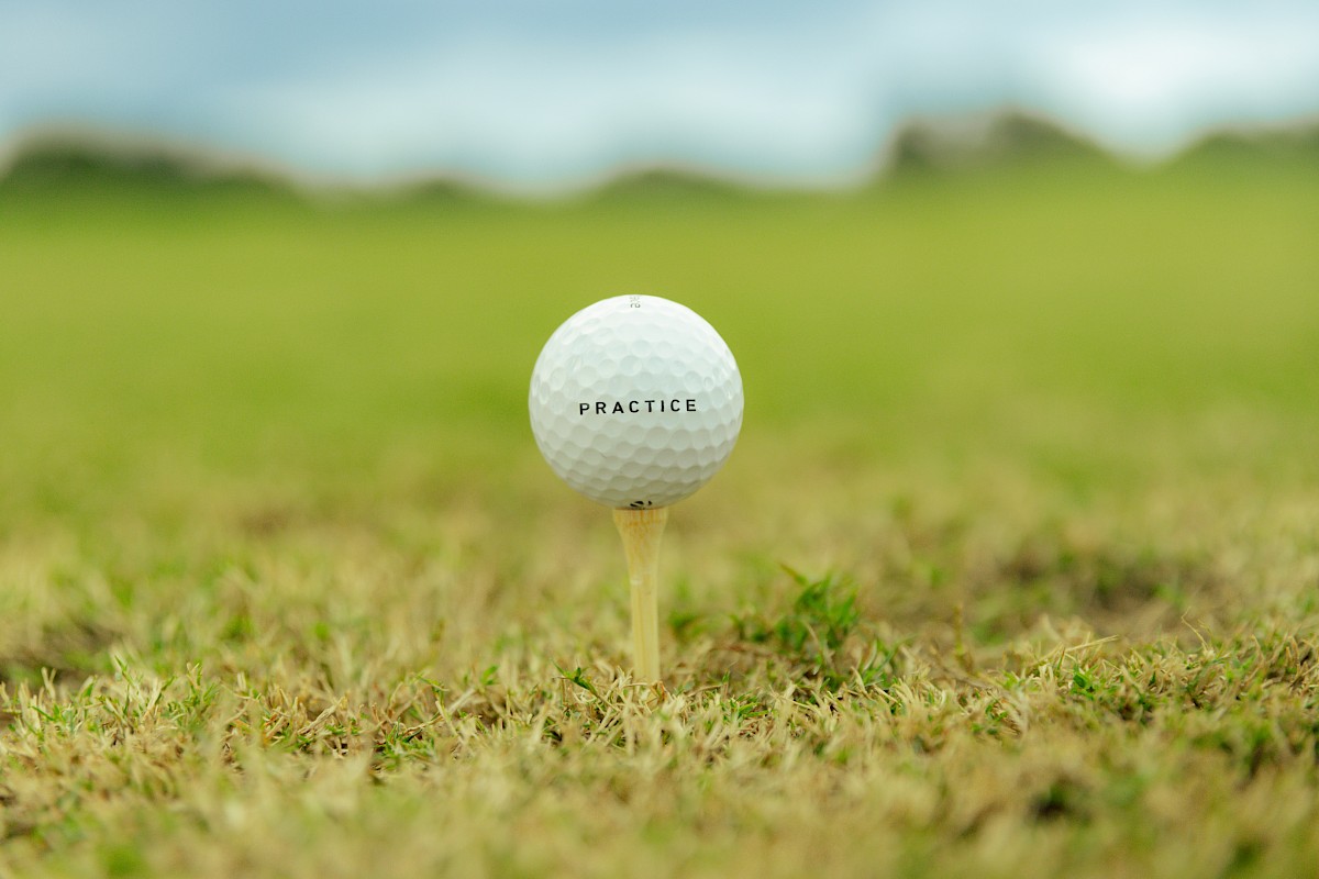 A white golf ball perched on a tee in a grassy field, with the word “PRACTICE” printed on it, ready for a swing under a blue sky.