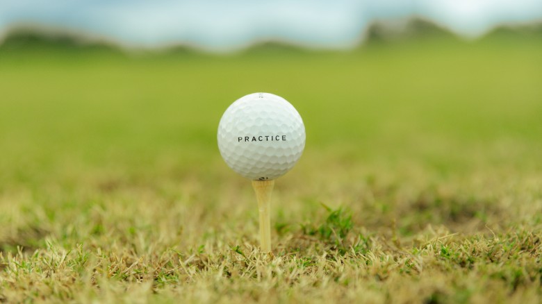 A white golf ball perched on a tee in a grassy field, with the word “PRACTICE” printed on it, ready for a swing under a blue sky.