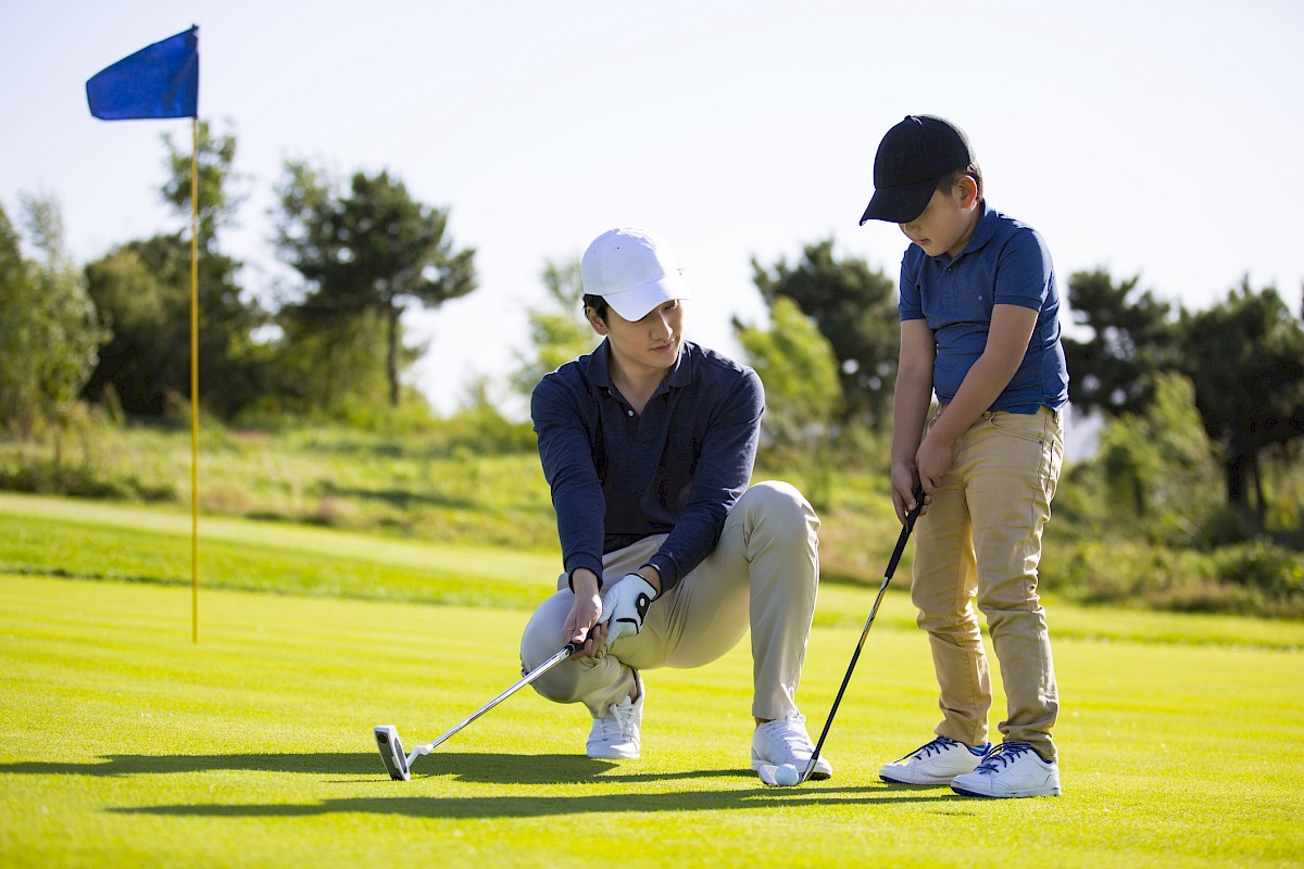 A person coaches a child in putting on a golf course, with a flag in the background and trees lining the horizon.