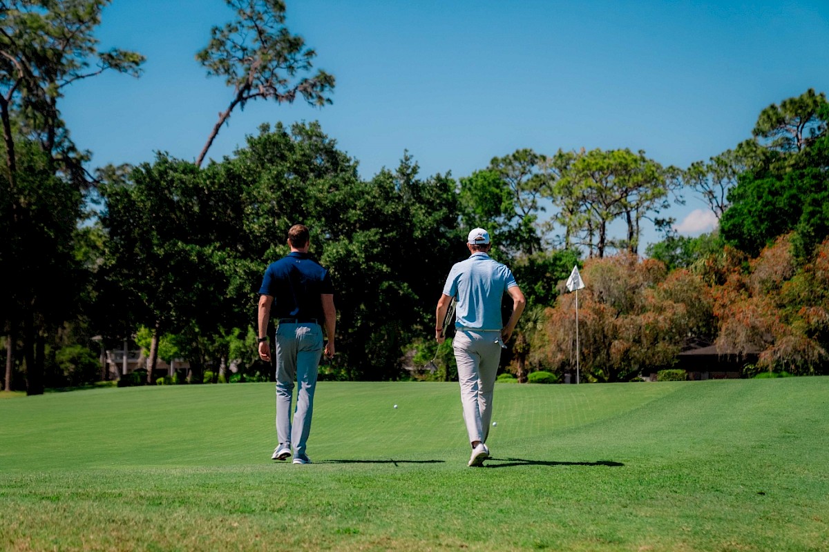 Two people walking on a golf course towards a flag, surrounded by trees and under a clear blue sky.