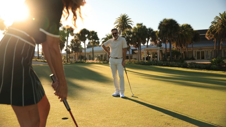 Two golfers on a sunny course: a woman in the foreground lining up a putt, a man watching with clubs, palm trees in the background, shadows long.