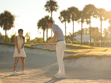 Two people play golf on a sandy course at sunset, with palm trees in the background and warm light.