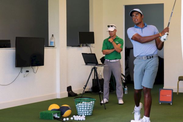 Two men in a golf simulator room, one swinging a club, the other watching
