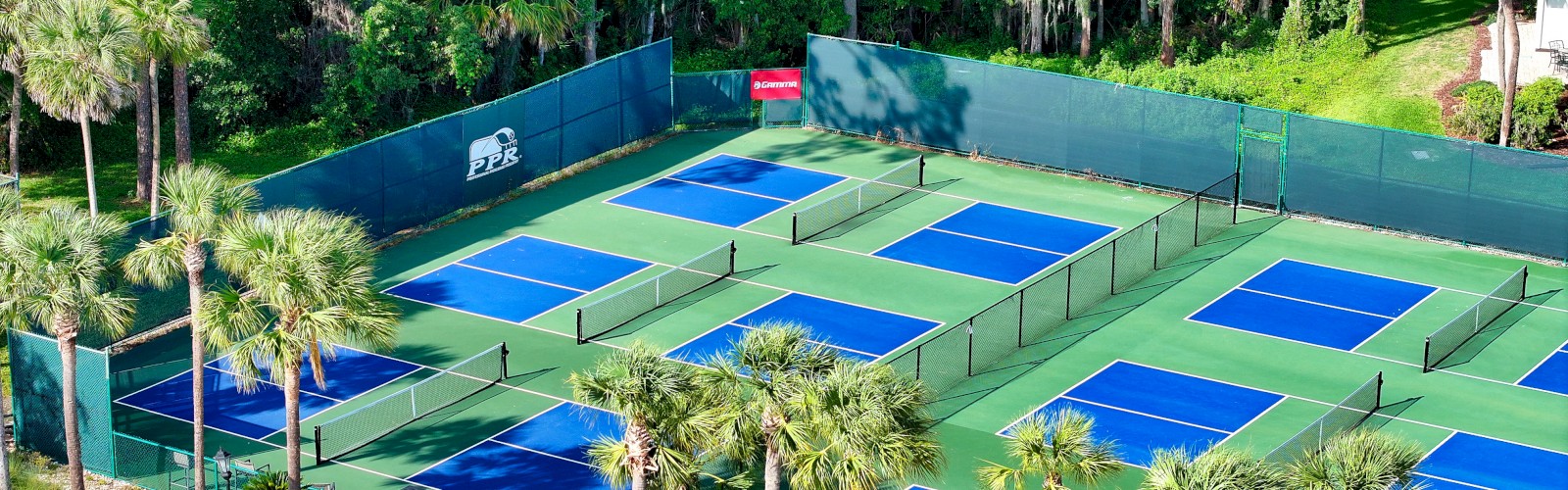 People playing on multiple blue tennis courts inside a fenced greenspace, surrounded by trees and palm trees, with sunny weather.