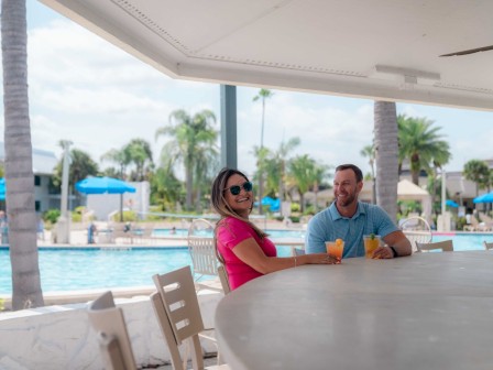 Two people are sitting at a poolside bar with drinks, enjoying the sunny weather.