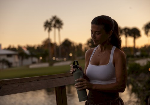 A fit woman in a white tank top stands on a wooden deck by the water at sunset, holding a spray bottle.
