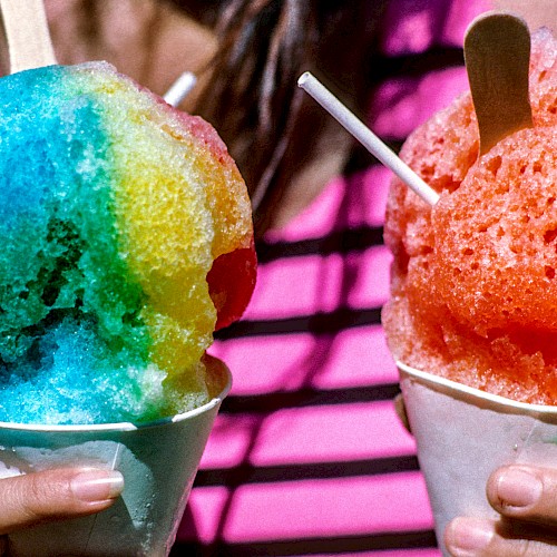 A person holds two colorful shaved ice treats in paper cups with wooden spoons, one blue and yellow, the other red, against a striped shirt background.