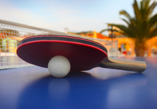 A ping pong paddle resting on a table tennis ball on a blue table with a net in the background and palm trees and buildings in the distance.
