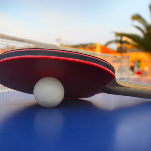 A ping pong paddle resting on a table tennis ball on a blue table with a net in the background and palm trees and buildings in the distance.