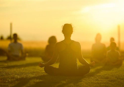 A group practices yoga outdoors at sunset, sitting on the grass in a meditative pose with a golden sky background.