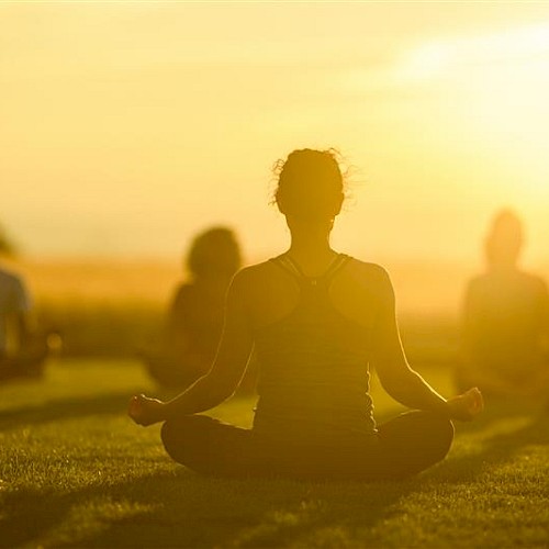 A group practices yoga outdoors at sunset, sitting on the grass in a meditative pose with a golden sky background.